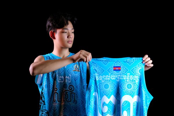 Young man displaying a vibrant sports jersey with intricate patterns and text on a black background in a studio setting.